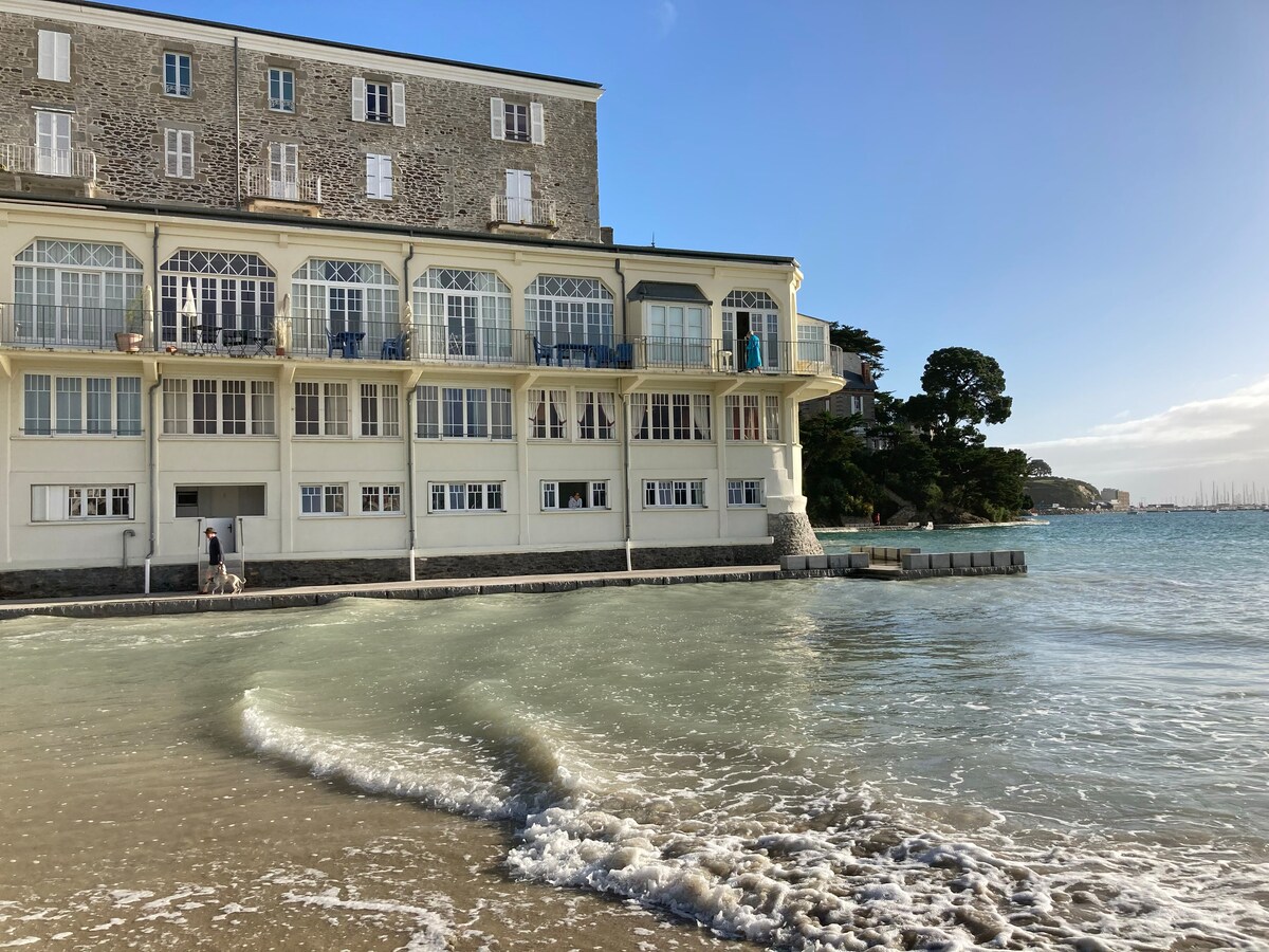 A beachfront building is captured from the water's edge, showcasing large windows and a prominent balcony on the upper level. The shoreline is visible, with gentle waves lapping against the sandy beach in the foreground, and distant greenery enhances the coastal scenery.