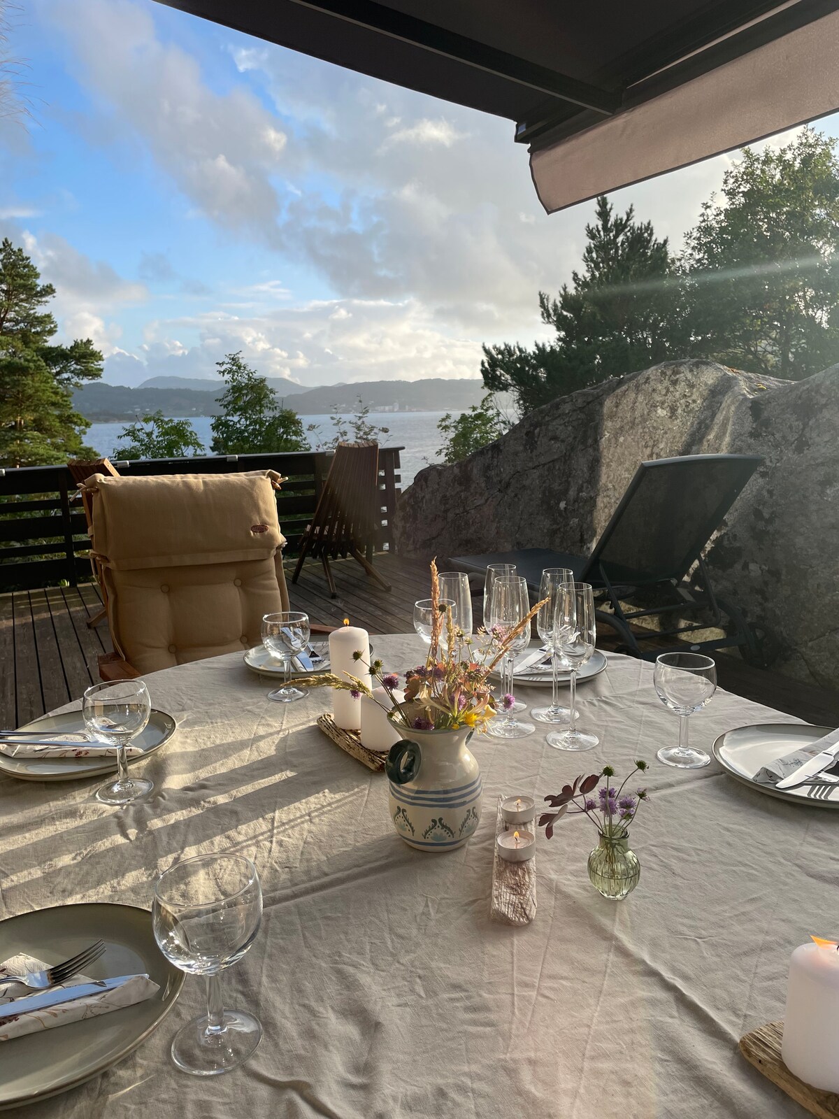 An outdoor dining setup is presented with a table covered by a light-colored cloth. Glassware and dishes are arranged neatly, accompanied by a delicate floral centerpiece. In the background, a scenic view of the water and surrounding trees is visible, enhancing the tranquil atmosphere.
