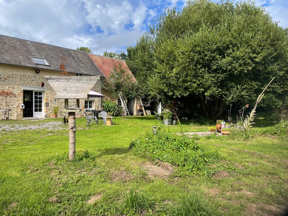 The image captures a natural garden setting surrounding a rustic stone building. Lush greenery and various plants fill the space, while a seating area with chairs and tables is visible. A pathway leads through the garden, inviting exploration and relaxation.
