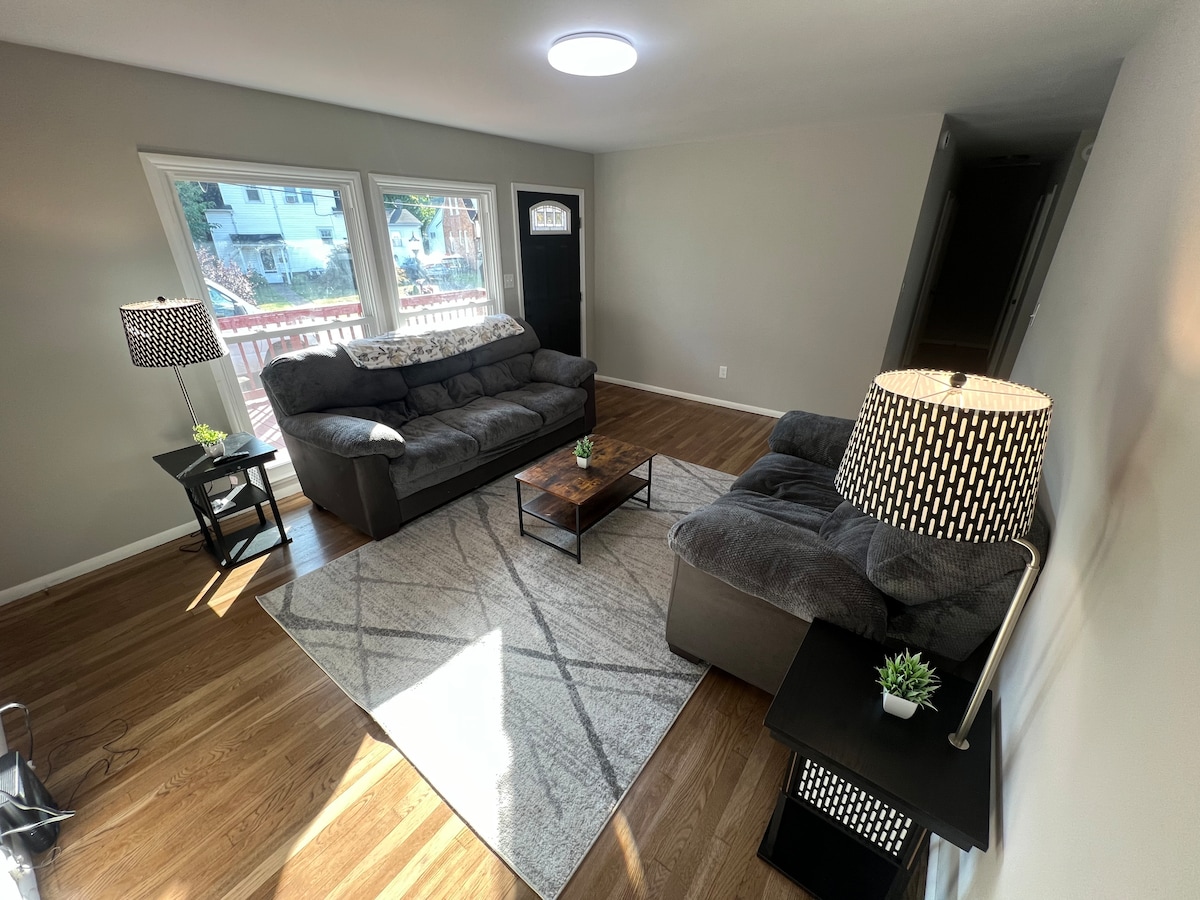 A welcoming living area features two upholstered sofas arranged around a coffee table. A patterned area rug covers the hardwood flooring, while large windows allow natural light to fill the space. Two side tables with lamps offer additional lighting and décor.