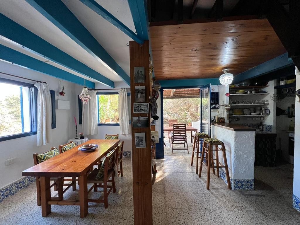 An open dining area features a large wooden table surrounded by chairs. Bright blue beams accent the ceiling, while natural light filters in through windows dressed with light curtains. A cozy seating area with wooden chairs and a bar is visible in the background.