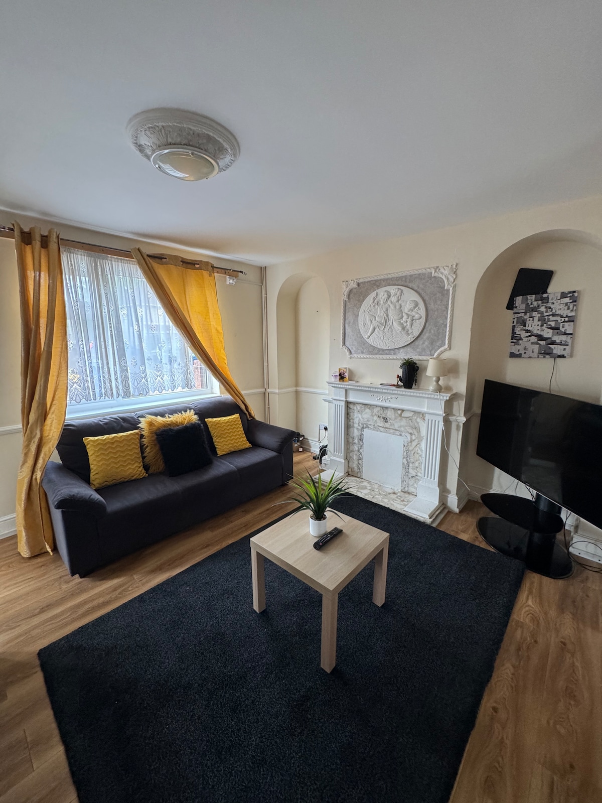 A cozy living area features a dark sofa adorned with yellow cushions, positioned near a small coffee table. A decorative fireplace adds character to the room, while large windows with sheer yellow curtains allow natural light to fill the space. A TV is mounted on the wall opposite.