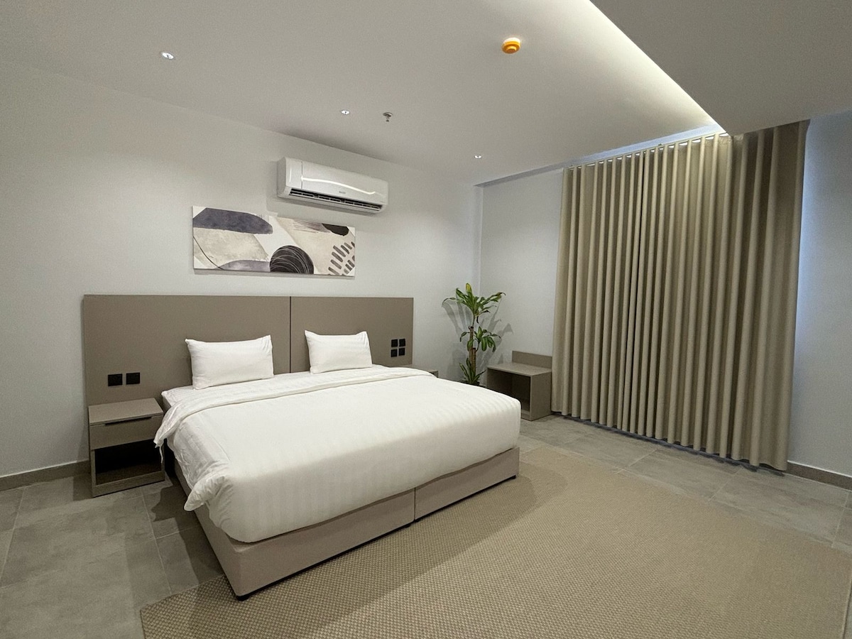 A spacious bedroom features a modern design with neutral tones. A large bed is centered against a minimalist headboard, complemented by two nightstands. Soft, light curtains frame the window, and a green plant adds a touch of color to the room.