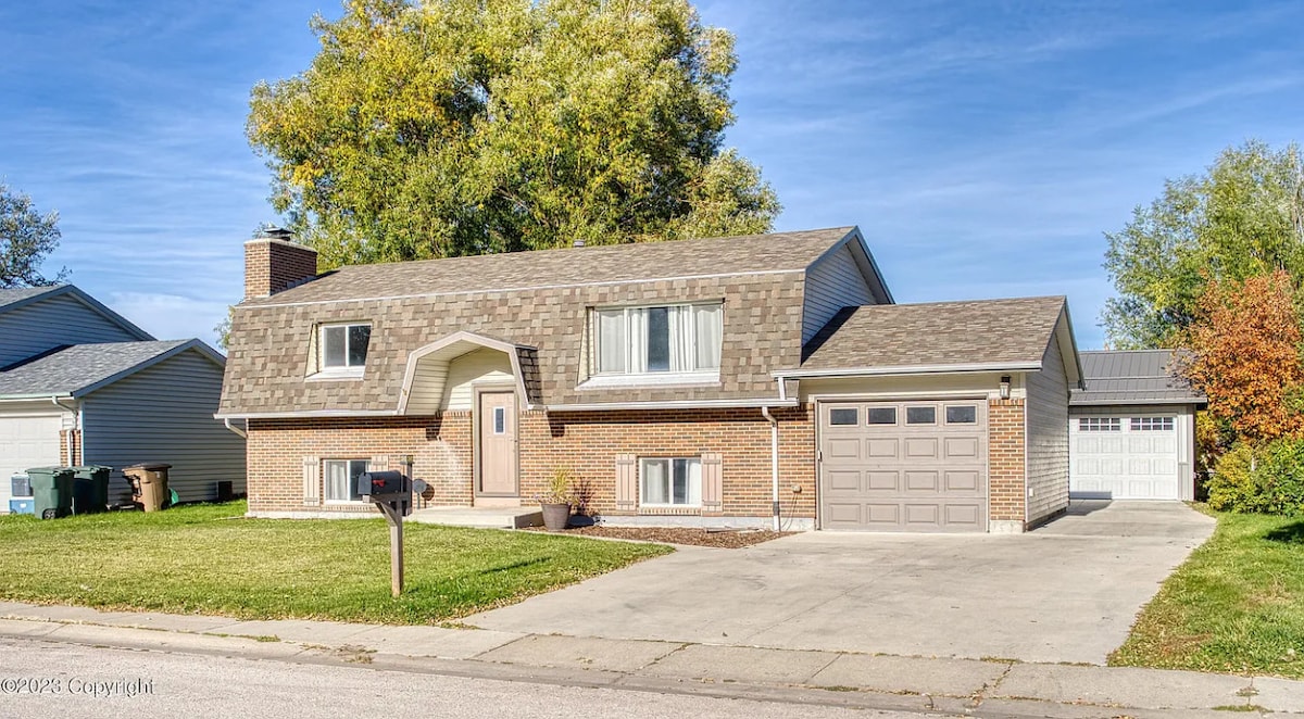 A two-story house with a peaked roof is situated on a green lawn. A gravel driveway leads to a two-car garage. Large windows allow natural light to enter, and trees provide a natural backdrop on either side of the property.