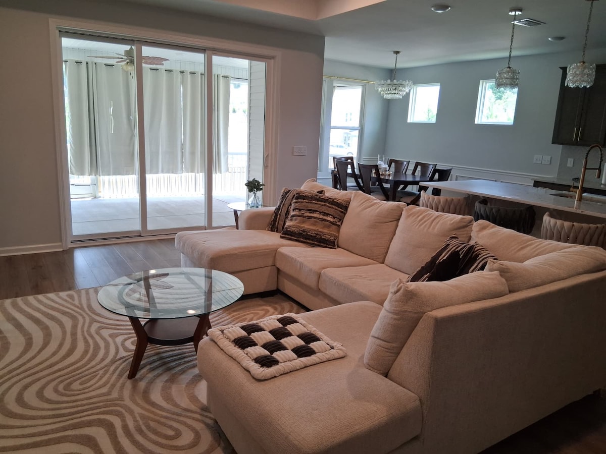 A spacious living room features a light-colored sectional sofa adorned with various throw pillows. A round glass coffee table rests atop a patterned area rug. Natural light flows through sliding glass doors, leading to an outdoor space, while a dining area is visible in the background.