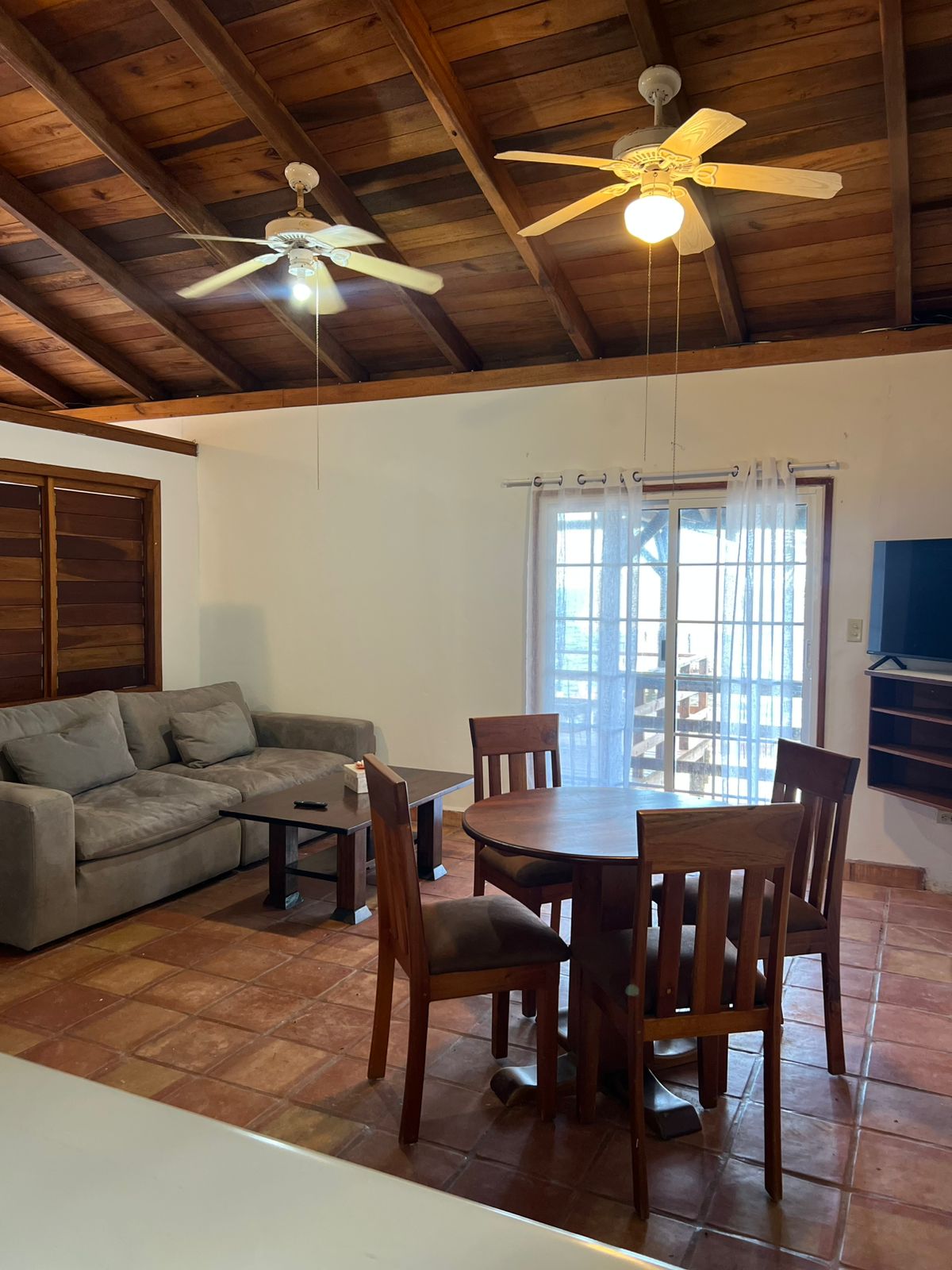 An open living space features a comfortable sofa and a round wooden dining table surrounded by chairs. Natural light enters through glass doors leading to the verandah, highlighting the high wooden ceiling and tiled floor.