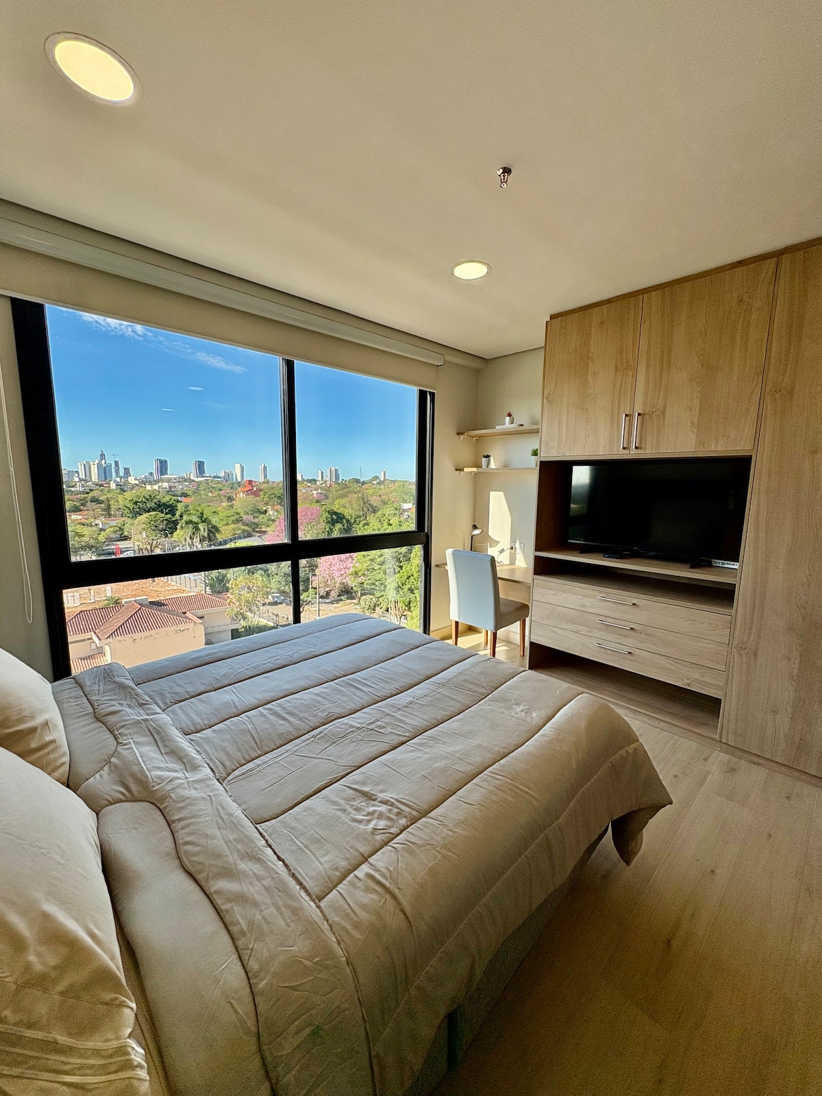 A well-lit bedroom features a spacious bed with a light-colored comforter, positioned next to a large window displaying a panoramic view of the city. Modern furnishings include a small desk and chair, and built-in cabinetry complements the warm wood tones.