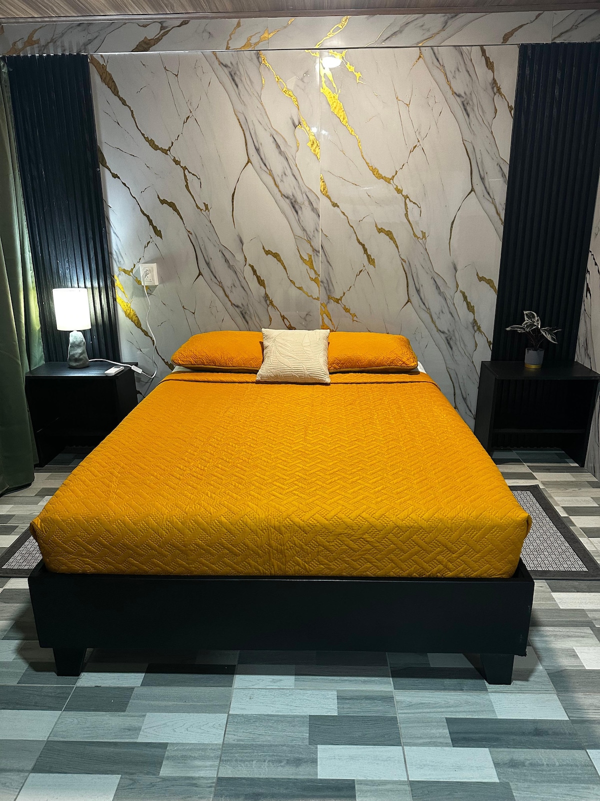 A modern bedroom features a spacious bed dressed in a vibrant yellow coverlet, complemented by a matching pillow. Black nightstands with lamps are positioned on either side. The walls exhibit a striking marble design with gold accents, and the floor consists of alternating tiles in varying shades.