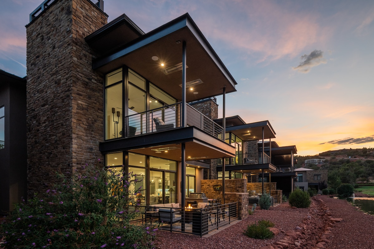 A modern townhome exterior is captured at dusk, featuring large glass windows and multiple balconies. The stone façade complements the evening sky, which is painted in soft hues. A gathering space with seating and a fire pit is positioned on the patio below.