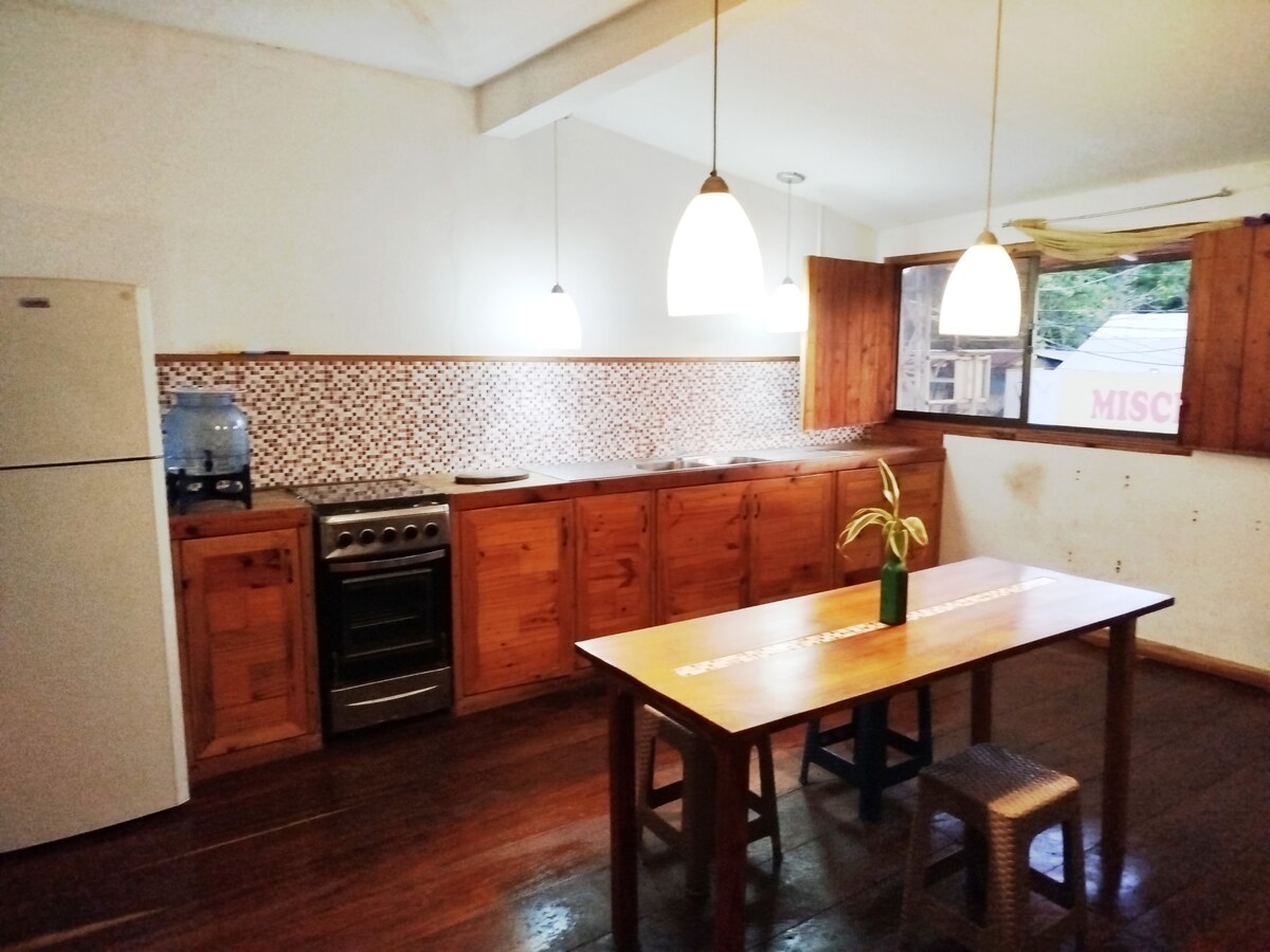 A well-equipped kitchen features wooden cabinetry and a spacious dining table. Bright pendant lights hang above, illuminating the space. A refrigerator and a stove are visible, with a mosaic backsplash adding texture to the area. Wooden floors contribute to a warm and inviting environment.