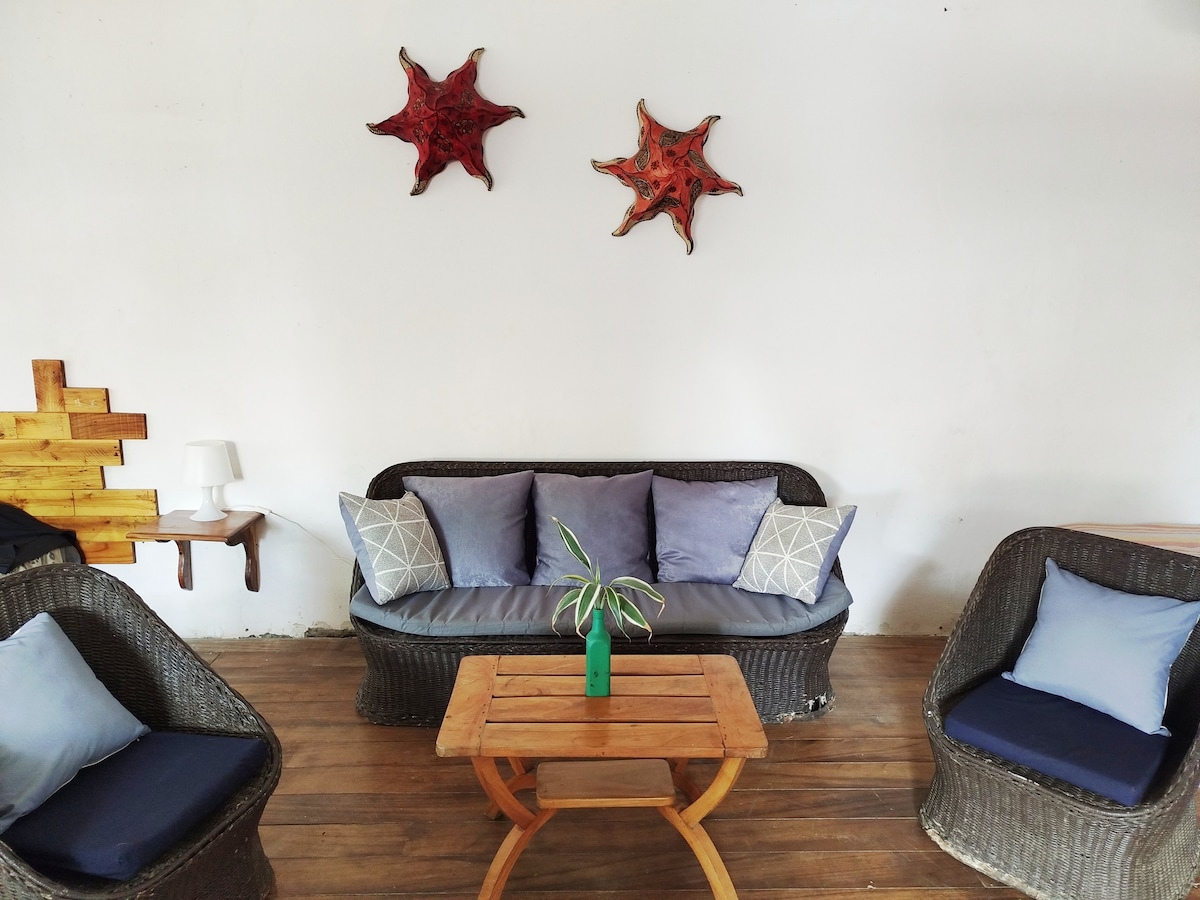 A seating area features a wicker sofa adorned with blue cushions, accompanied by two matching armchairs. A wooden coffee table is positioned at the center, with a small green vase placed on top, and two decorative star-shaped wall pieces are displayed above the sofa.