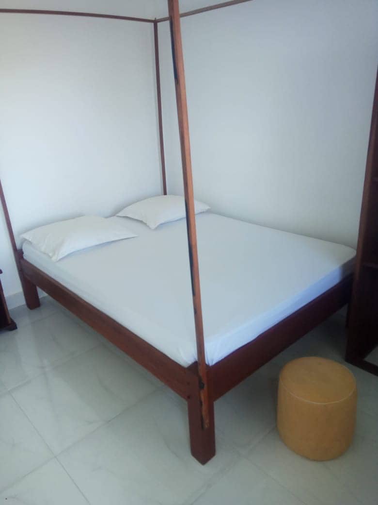 A spacious bed with a simple wooden frame is situated against a plain wall. Two pillows rest neatly on the white linens. A small round stool made of light-colored material is positioned nearby, adding to the uncluttered space.