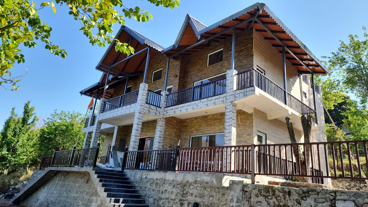 A modern villa is positioned on a gently sloping hillside, featuring a mix of stone and wooden finishes. Two levels are visible, each with spacious balconies that overlook the surrounding greenery, framed by tall trees under a bright blue sky.