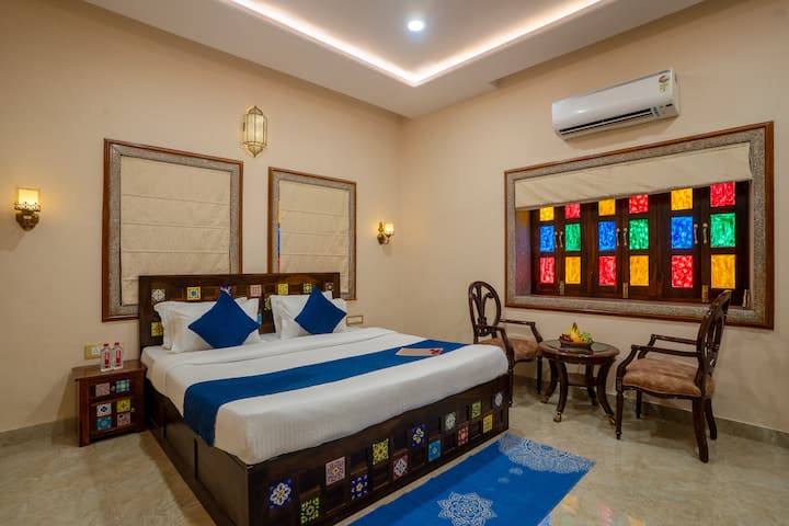 Bedroom 2 – Neel Mahal: Elegant and serene, featuring a double bed with blue accents, colorful tile headboard, stained glass window, cozy seating with fruit basket, ambient lighting, and AC for comfort. A perfect blend of charm and calm.
