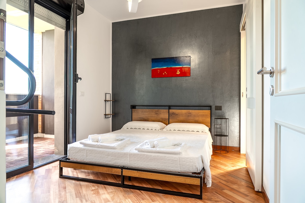 A well-appointed sleeping area features a spacious bed with neatly arranged towels. A striking wall art piece provides a pop of color against the contrasting grey wall. Large glass doors offer access to an outdoor terrace, enhancing the natural light within the room.