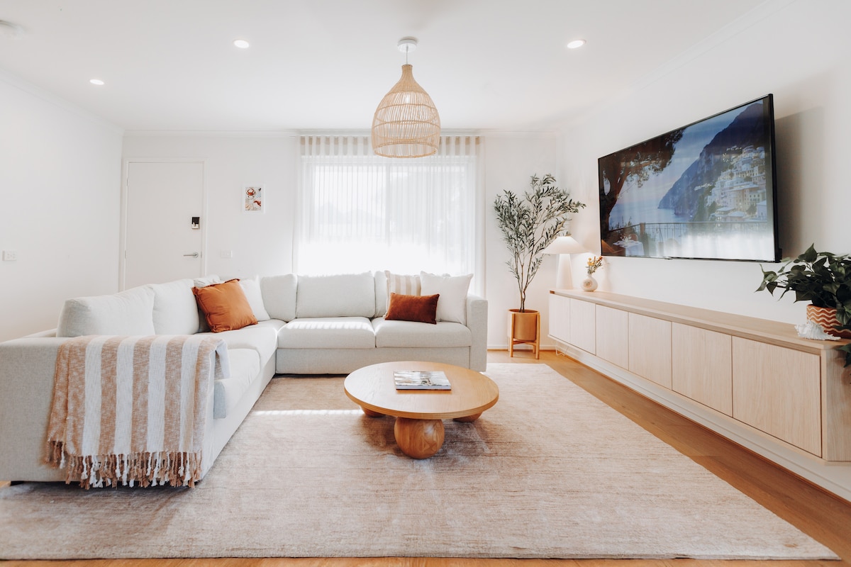 A spacious living area is highlighted by a large, light-colored couch adorned with a striped throw. A circular wooden coffee table centers the room, which is anchored by a plush area rug. Above, a wall-mounted television displays soothing scenery, with natural light filtering through sheer curtains.