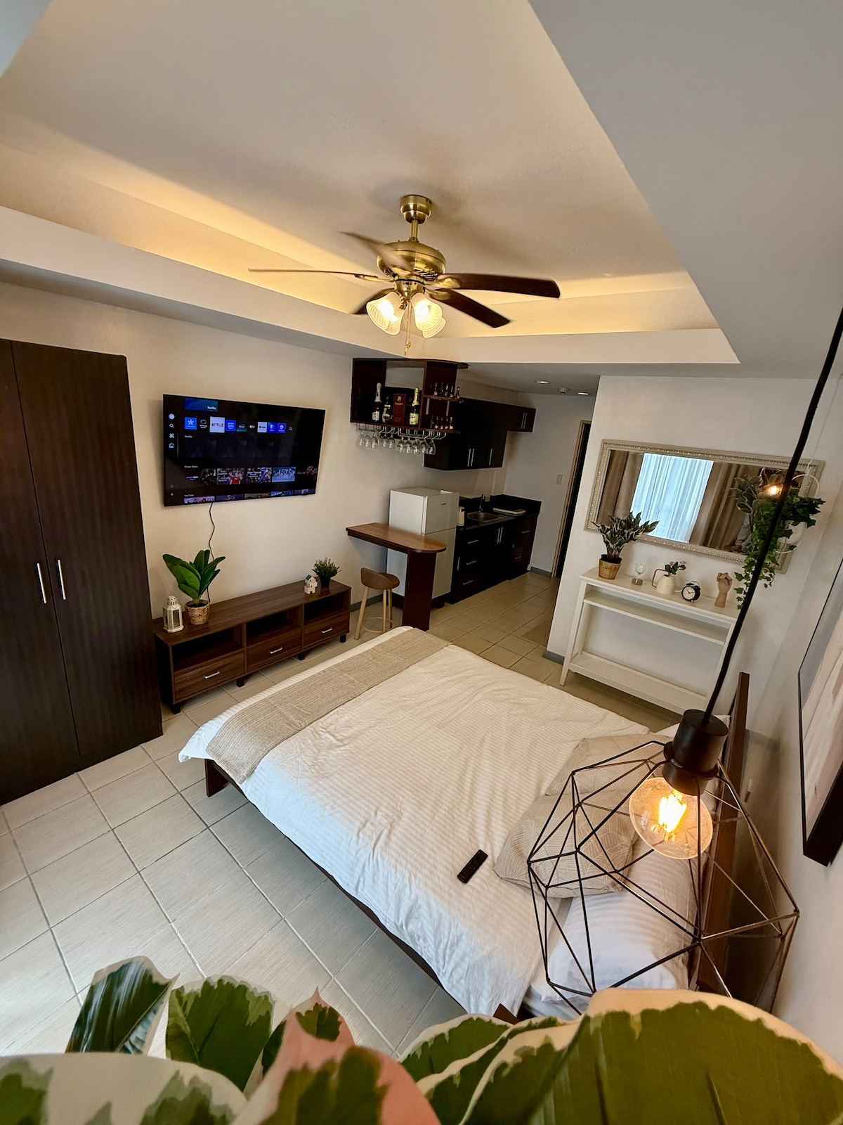 A well-organized studio space features a spacious bed with white linens, complemented by a decorative pendant light. A wall-mounted television is visible alongside a kitchenette with essential appliances. A large mirror and plants add an element of greenery, while soft lighting enhances the room’s ambiance.
