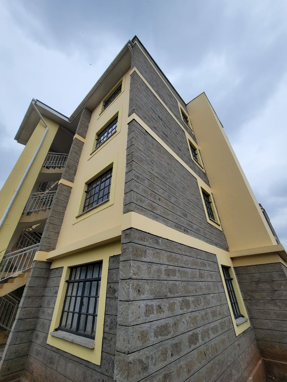 The exterior of a three-story apartment building is showcased, featuring a mix of grey stone and yellow-painted walls. Large windows with black grilles are visible on each floor, enhancing natural light. A staircase can be seen on the side, providing access to individual units.