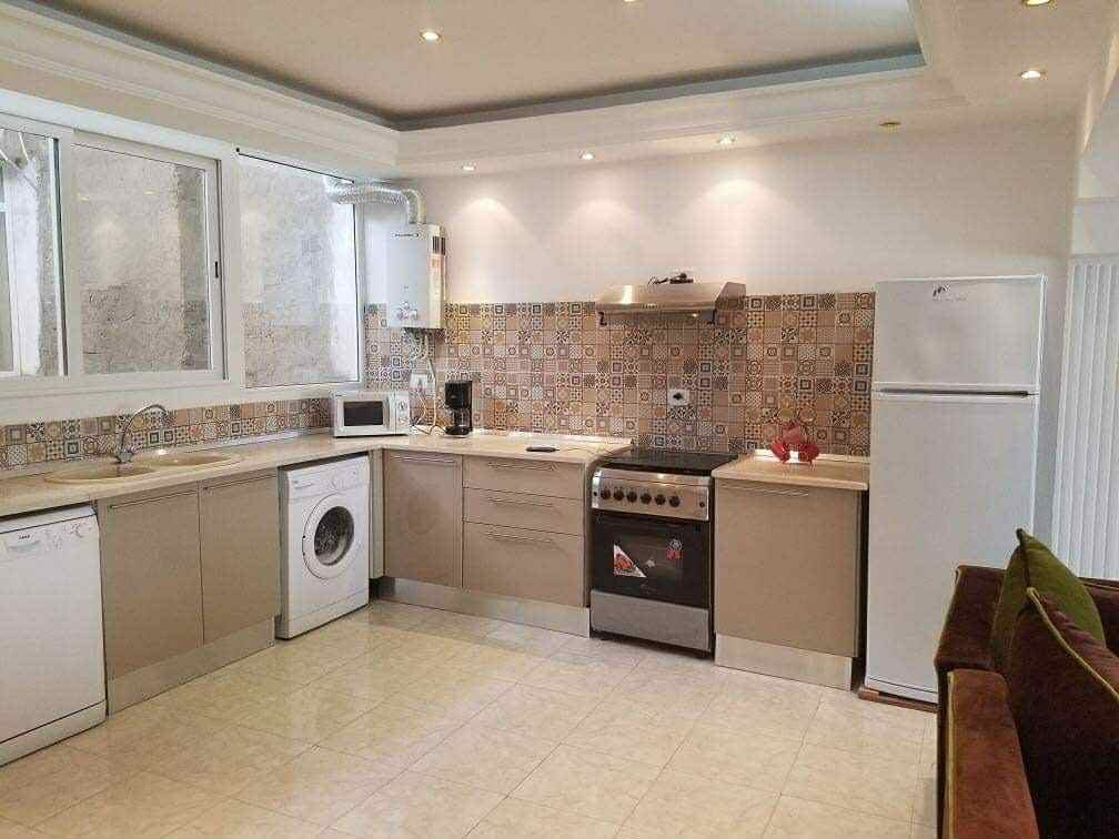 A modern kitchen is presented with beige cabinetry and a patterned tiled backsplash. Stainless steel appliances, including a stove, microwave, and refrigerator, are visible. A washing machine is integrated into the space, which is illuminated by natural light coming through multiple windows.