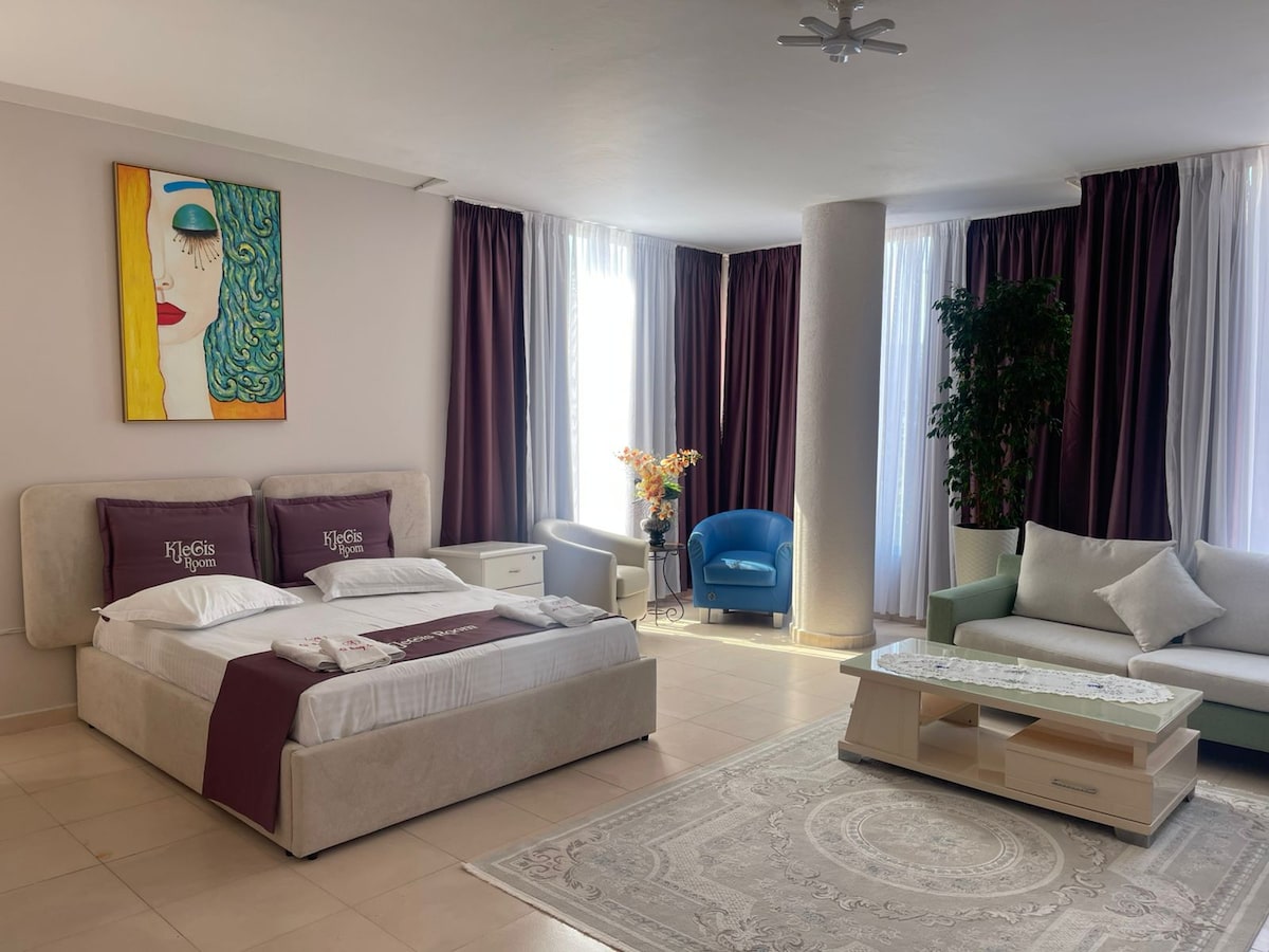 A spacious sleeping area is showcased with a neatly made bed featuring white linens. A vibrant artwork adorns the wall above. A comfortable blue chair and a light-colored sofa are positioned nearby, complemented by large windows allowing natural light to fill the room.