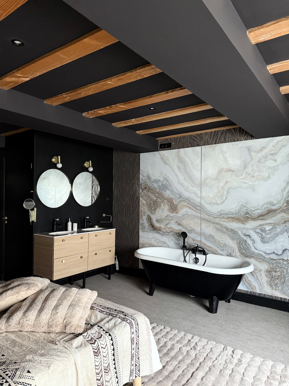 A modern bathroom features a freestanding black and white bathtub positioned against a marbled stone wall. A wooden vanity with dual sinks is flanked by circular mirrors, creating a balanced look. Recessed lighting adds subtle illumination to the space.