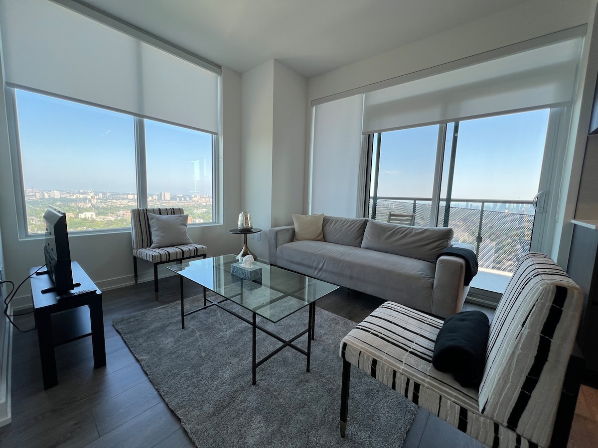 A spacious living area is highlighted by floor-to-ceiling windows offering expansive city views. A light-colored sofa and two patterned armchairs surround a modern glass coffee table. Natural light fills the room, enhancing the neutral decor and subtle textures of the furnishings.