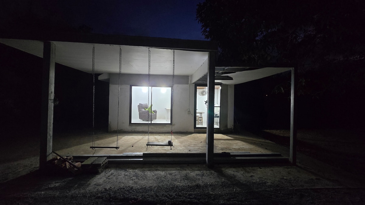 An outdoor space is illuminated softly by light from a large window, revealing a simple interior with a chair and plant. Two swings hang on a concrete patio, surrounded by a tranquil evening atmosphere.