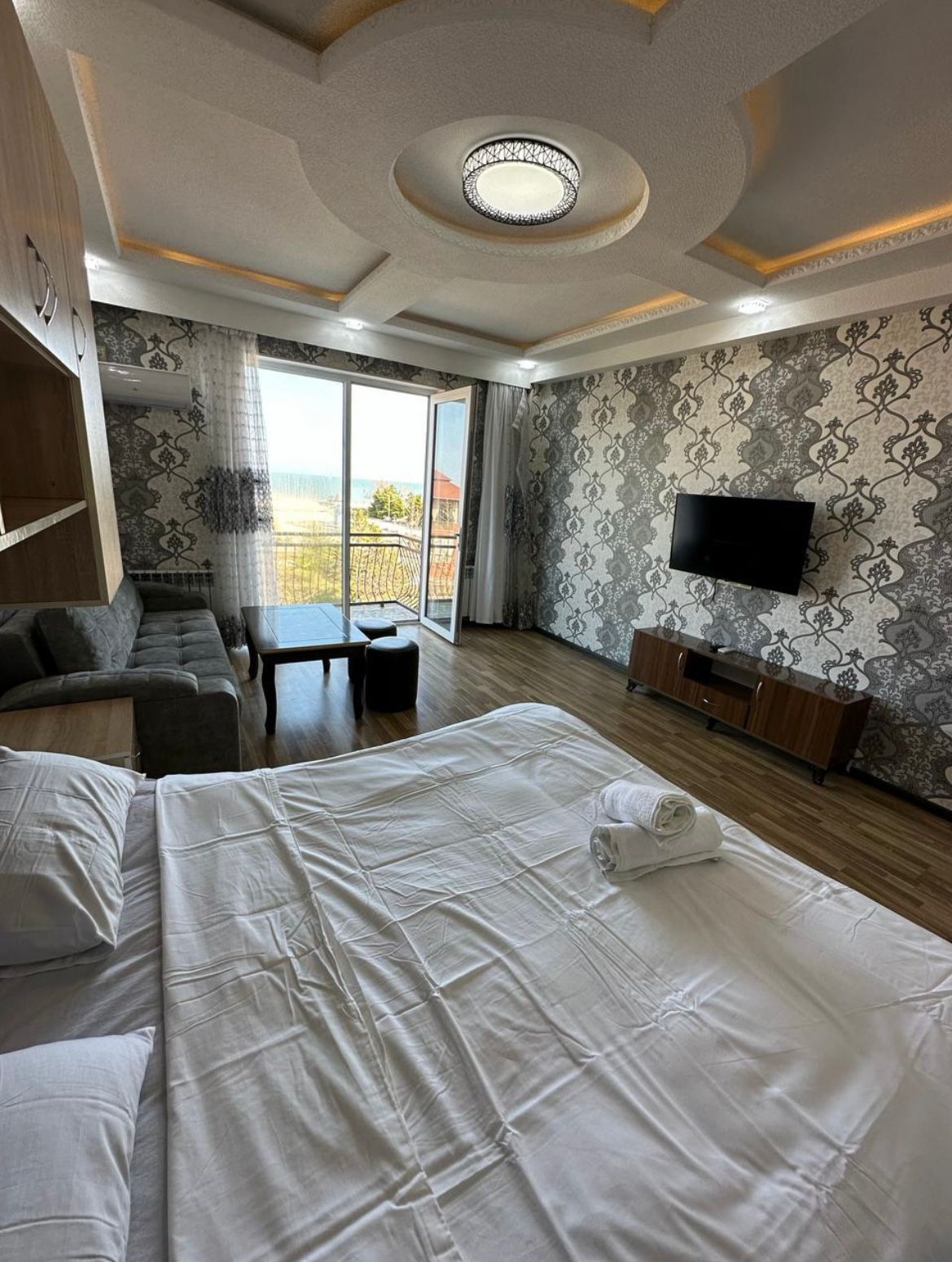 A spacious bedroom features a large bed with white linens and rolled towels. Natural light floods the room through floor-to-ceiling glass doors leading to a view of the sea. A comfortable seating area with a gray sofa is visible, along with a flat-screen television mounted on the wall.