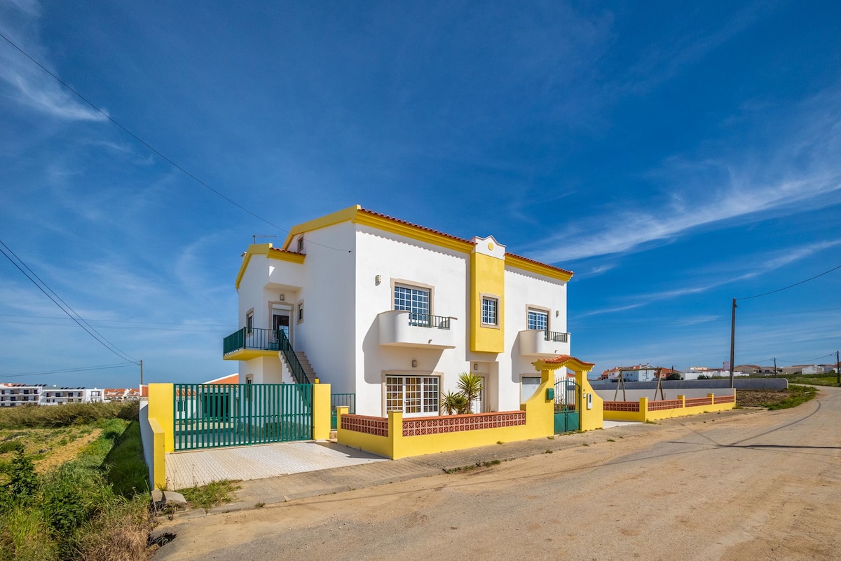 A spacious detached villa is depicted, featuring a bright yellow and white exterior. The property includes multiple windows and a large entrance gate. A welcoming pathway leads to the entrance, highlighted by landscaped greenery and a clear blue sky in the background.