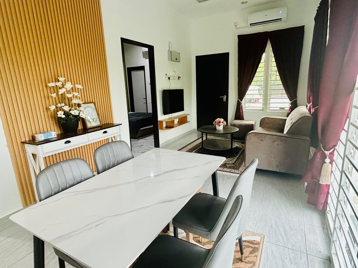 A modern dining area features a sleek marble-top table surrounded by four chairs. A comfortable sofa is seen in the background, complemented by large windows dressed in burgundy curtains. Decorative plants add a touch of greenery, while a wall-mounted television and a framed mirror enhance the space.