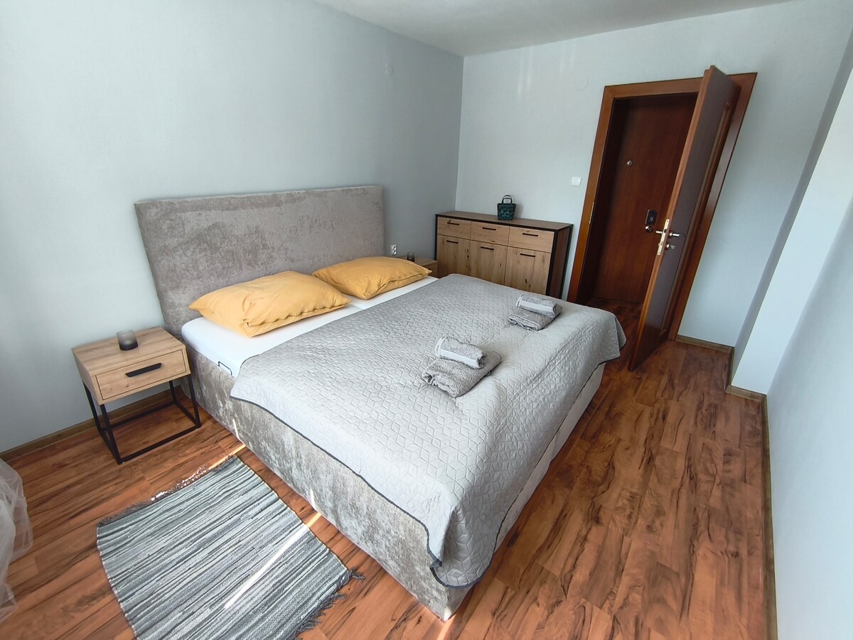 A comfortable sleeping area features a soft bed with neutral bedding and two bright yellow pillows. A small nightstand is positioned beside the bed, and a textured rug lies on the wooden floor. An open door leads to another room, enhancing accessibility.