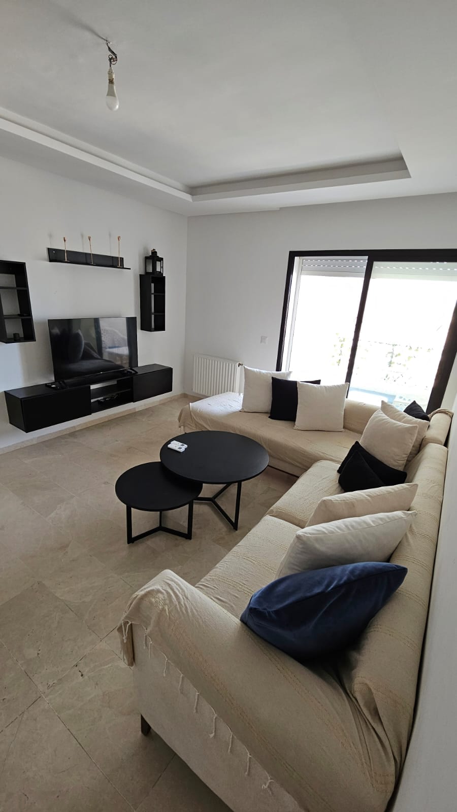 A light-filled living area features a beige sofa adorned with dark cushions, paired with two black coffee tables. A modern television is mounted on the wall, accompanied by minimalist shelving units. Large windows offer views of the outside, creating an inviting atmosphere.