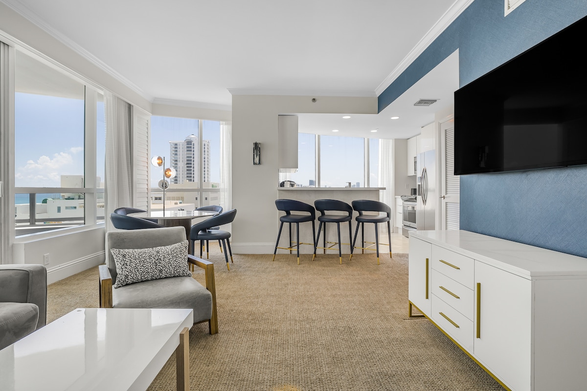 The elegant living area is highlighted by large windows providing ample natural light, featuring a modern sofa and a spacious table surrounded by four stylish chairs. A flat-screen TV is mounted on the wall, and a fully equipped kitchen area is visible in the background.