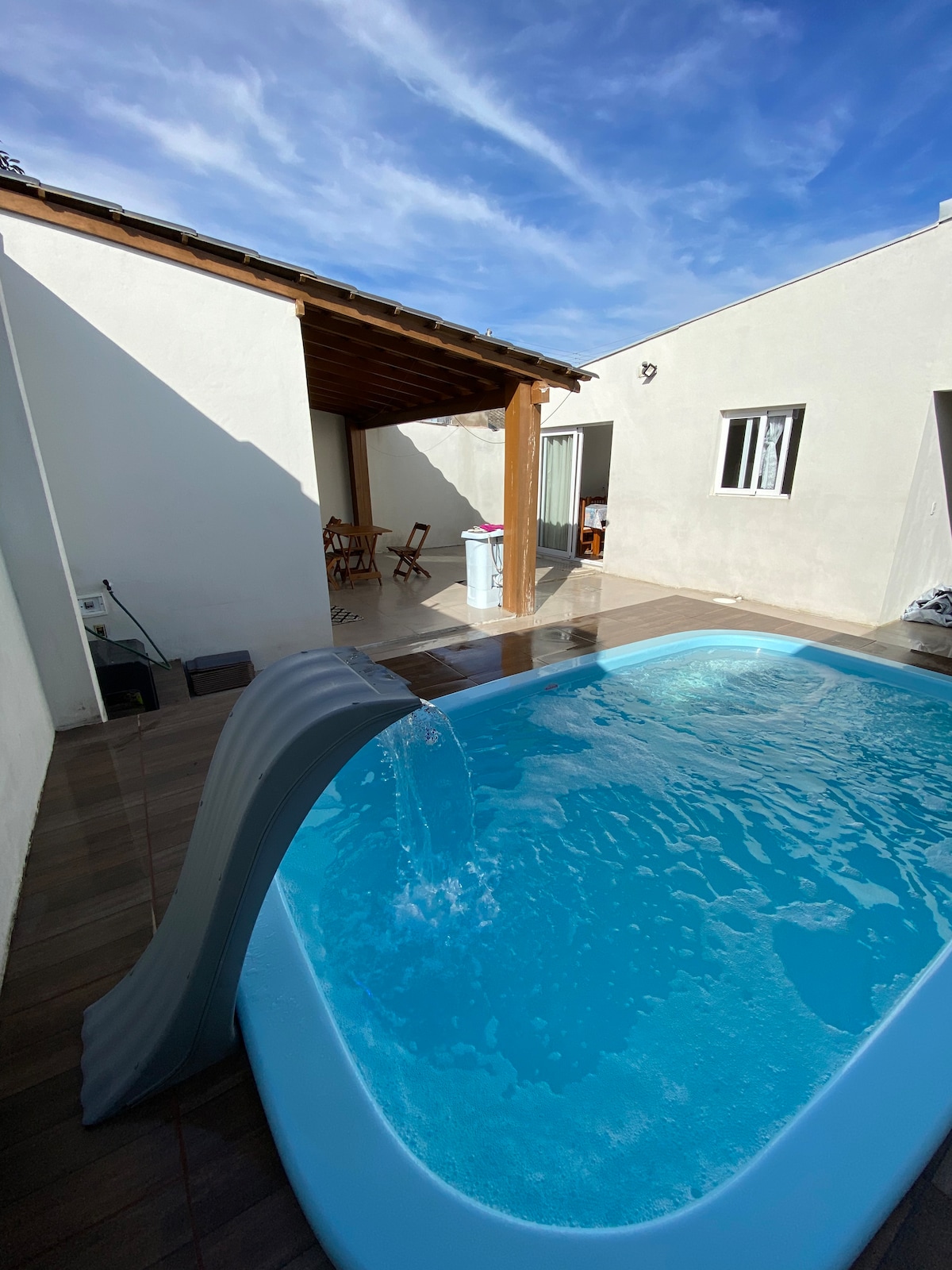 A swimming pool is visible in the foreground, featuring a slide on one side. Behind the pool, an outdoor seating area with a wooden table and chairs provides a space for relaxation. The area is surrounded by white walls and a roof structure, under a clear blue sky.