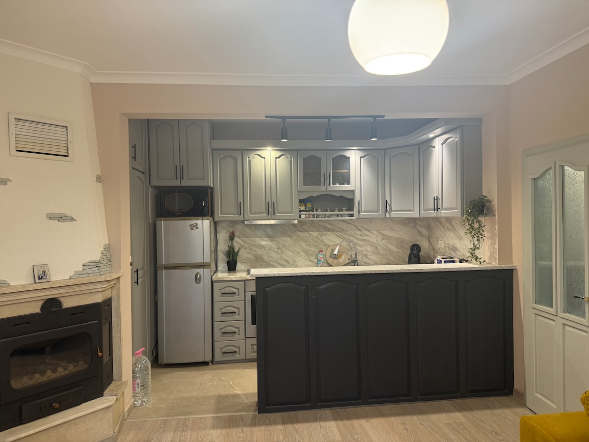 A modern kitchen area features grey cabinetry with a marble-patterned backsplash. A silver refrigerator stands against the cabinetry, while a small plant adds a touch of greenery. The kitchen space is well-lit by overhead fixtures, creating a functional cooking environment.