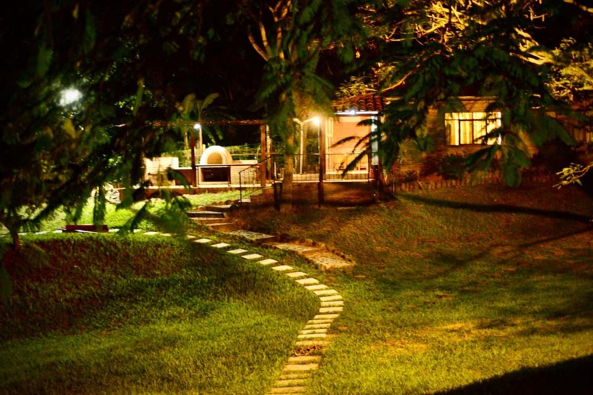 A serene pathway of stepping stones winds through the lush, green yard, leading towards a well-lit outdoor area. Soft illumination highlights nearby trees and the inviting ambiance of the surrounding space, creating a peaceful setting ideal for relaxation.