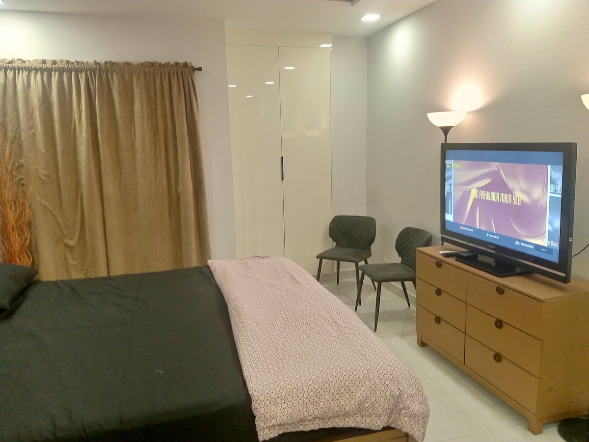 A neutral-colored sleeping area features a spacious bed with a textured blanket. Two chairs are positioned against the wall, and a television stands on a wooden dresser. A curtain provides privacy, and soft lighting enhances the cozy atmosphere.
