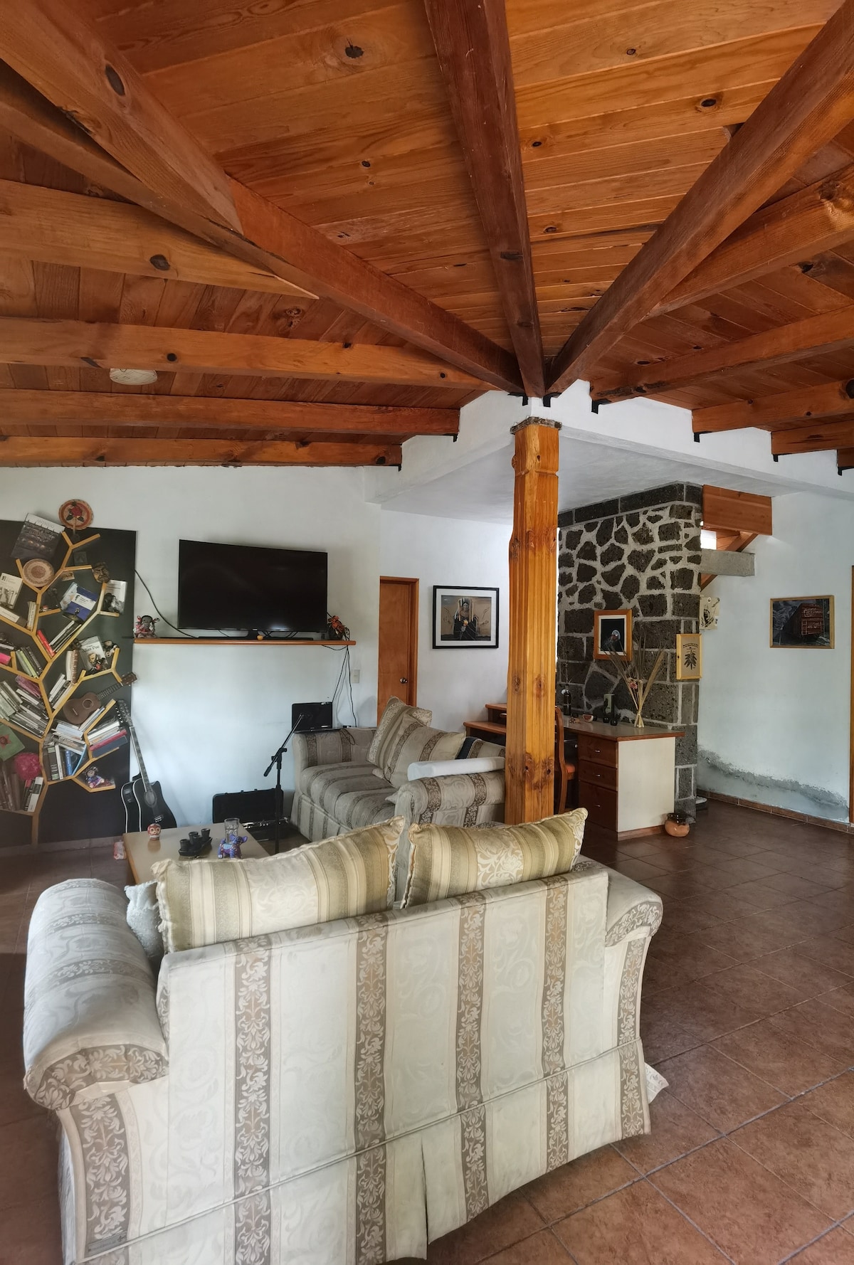 A cozy living space features wooden beams overhead and a stone accent wall. Two comfortable armchairs are positioned toward a television mounted on the wall. Decorative elements and artwork are subtly integrated, contributing to a warm environment.
