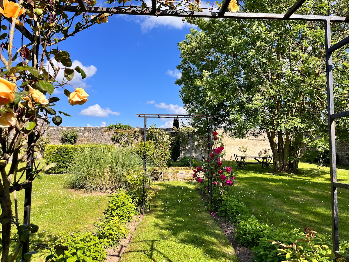 A spacious garden featuring vibrant greenery and colorful flowers, with a wooden pergola in the background. Seating areas are visible, inviting relaxation in a serene outdoor setting. The blue sky and scattered clouds create a peaceful atmosphere, surrounded by lush plants and well-maintained lawn.