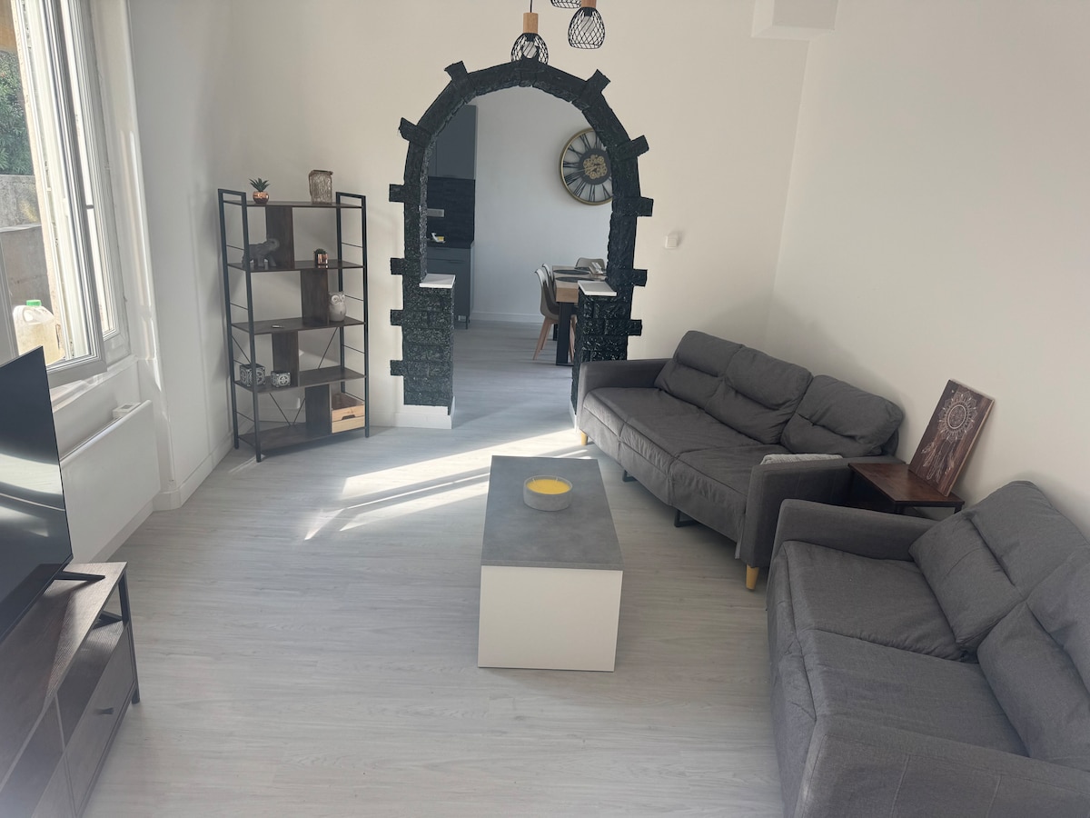 A bright living room is showcased, featuring two gray couches arranged around a square coffee table. A decorative archway leads to the dining area. Natural light streams in through the windows, highlighting a sleek shelving unit and a wall clock.