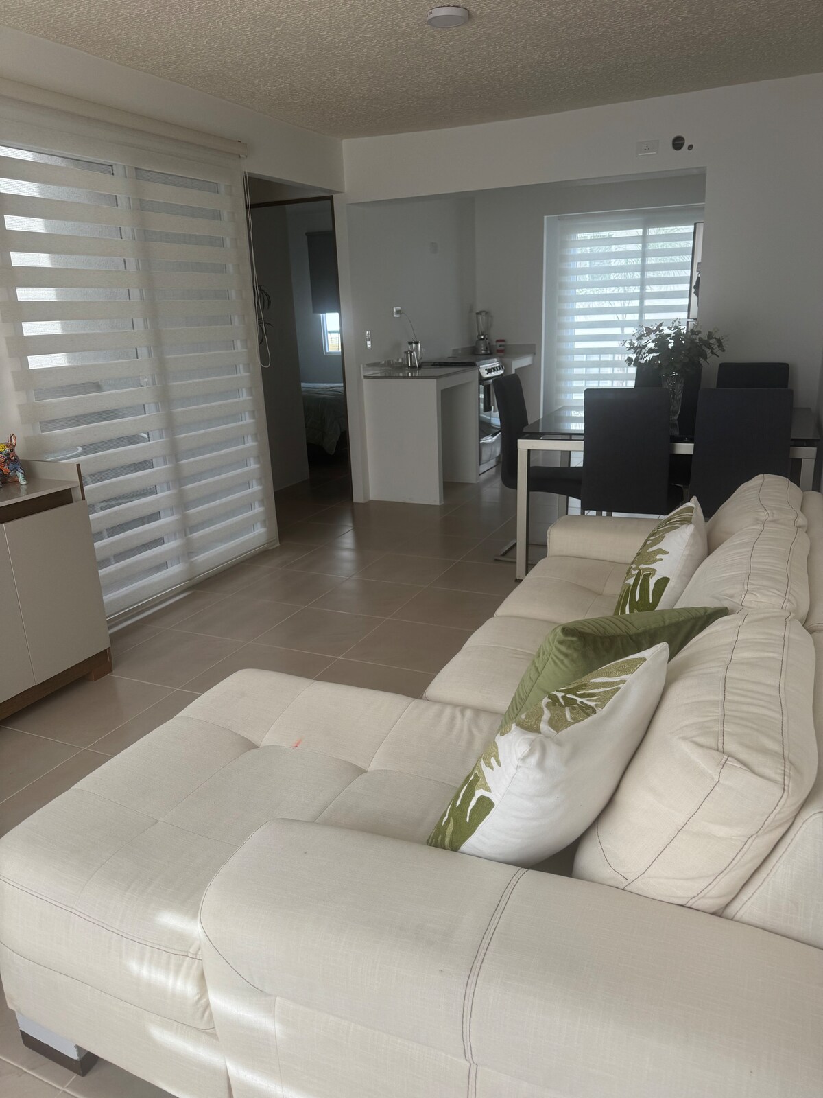 A modern living space is highlighted by a light-colored sectional sofa, adorned with decorative pillows. A dining area with dark chairs is visible in the background, alongside large windows allowing diffused light to enter, while sleek blinds add privacy.