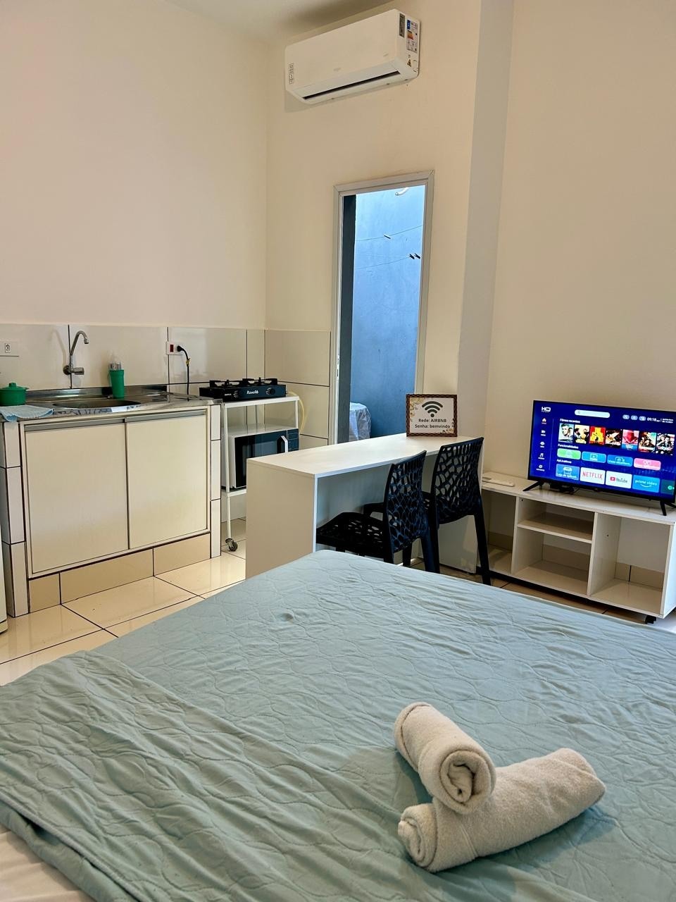 A modern kitnet features a spacious bed with a light blue coverlet. A kitchenette with essential appliances is visible, alongside a small dining table with two black chairs. A television is positioned on a stand, and a window allows natural light into the room.