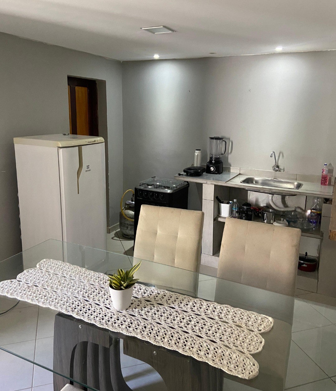 A modern kitchen area is visible, featuring a glass dining table with two beige chairs. A countertop is equipped with a sink, a stove, and a blender. A refrigerator can be seen to the left, and natural light illuminates the space.