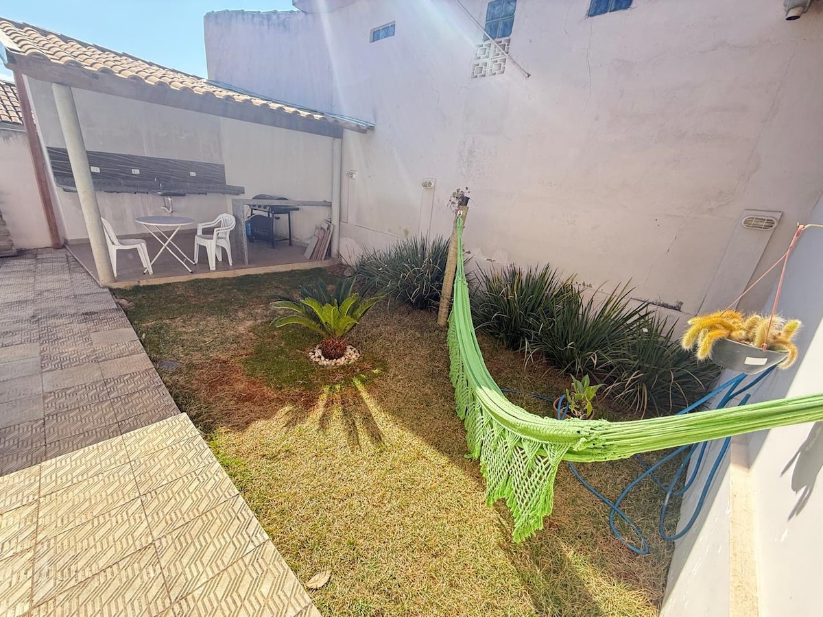 A sunny outdoor area is depicted with a green hammock gently swaying. Lush grass and low shrubbery frame a small garden space. A covered area features a table and chairs, ideal for dining or socializing. The textured stone flooring provides a warm and inviting feel.