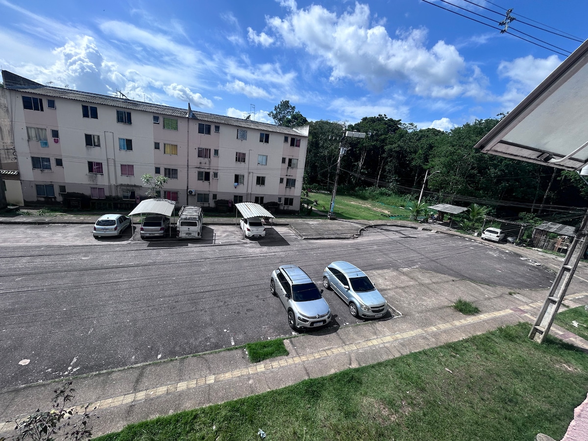 A view from the accommodation shows a spacious parking area in front of multi-story buildings. Several vehicles are parked, with green trees visible in the background, offering a serene touch to the surroundings.