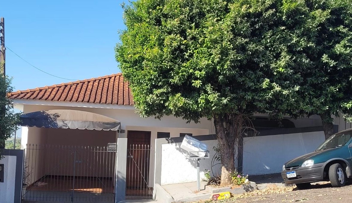 The exterior of the house features a white facade with a brown tiled roof, framed by a large green tree. A gated entrance provides access to the property, which includes a modest driveway with a parked car. Bright blue sky can be seen overhead.