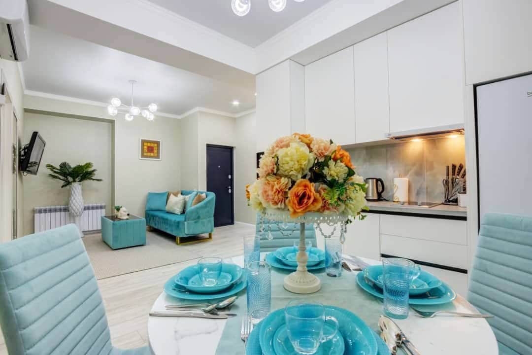 A bright dining area features a marble table set with turquoise plates and glasses. A vase with flowers in soft hues adds a decorative touch. The backdrop shows a spacious living area with a light teal couch and a flat-screen TV, enhancing the room's modern design.