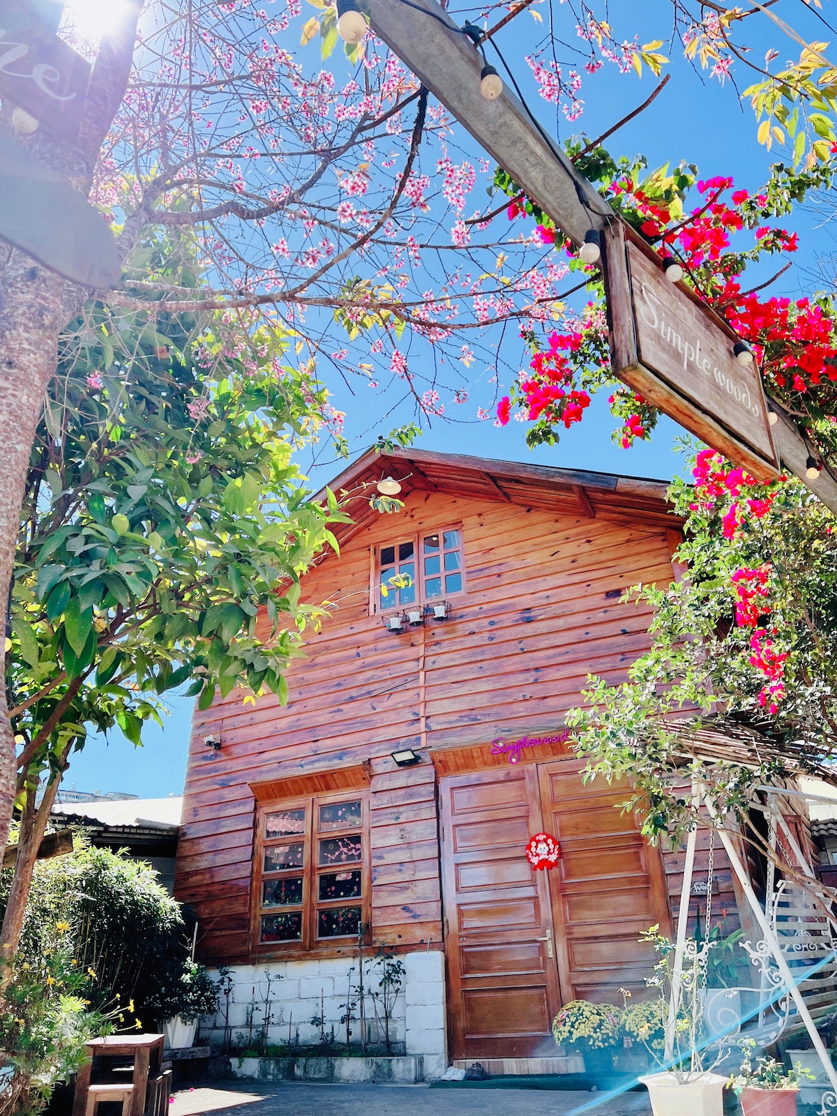 A wooden house characterized by a rustic design is surrounded by greenery and blooming pink flowers. Bright blue skies create a vibrant backdrop, while a sign hangs nearby, contributing to the welcoming atmosphere of the entrance.