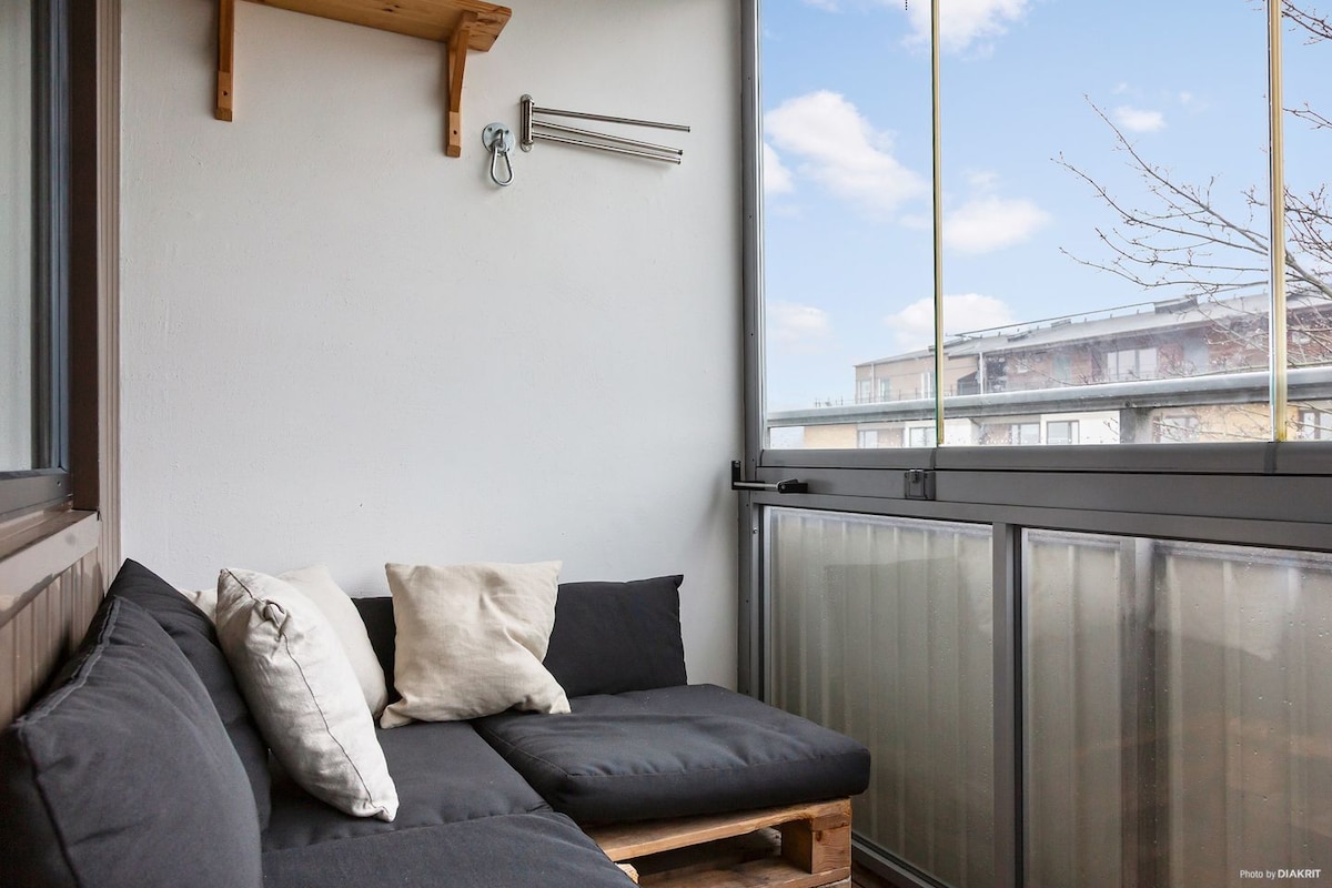 A cozy balcony area features a seating arrangement made of dark cushions placed on a wood-framed base. Bright light enters through large glass windows, creating a welcoming space to relax and enjoy the view. A small shelf is mounted on the wall for added convenience.