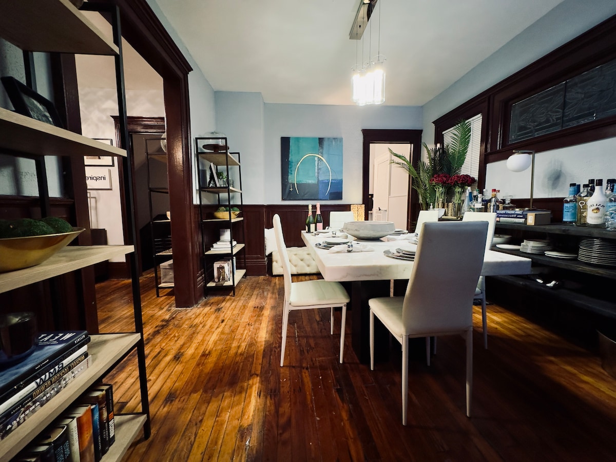 A dining area features a long, rectangular table set for a meal, surrounded by white chairs. Natural wood flooring enhances the space. A painting with abstract shapes hangs on the wall, while shelves display various books and decorative items, providing an organized atmosphere.